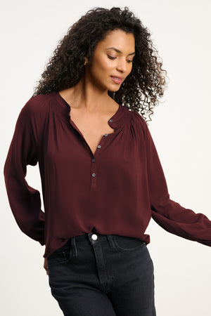 A woman wears the AURELIA TOP by Velvet by Graham & Spencer—a long-sleeve, burgundy blouse with smocked details and button accents—paired with black jeans. The relaxed rayon challis fit ensures comfort against a plain light background.