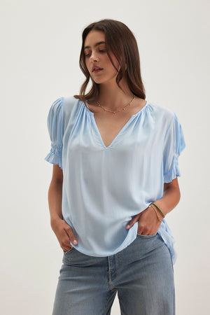 A person with long hair stands wearing a Velvet by Graham & Spencer CALISSA SPLIT NECK BLOUSE made from soft rayon challis, paired with jeans. Their hands rest in their pockets against a plain background.