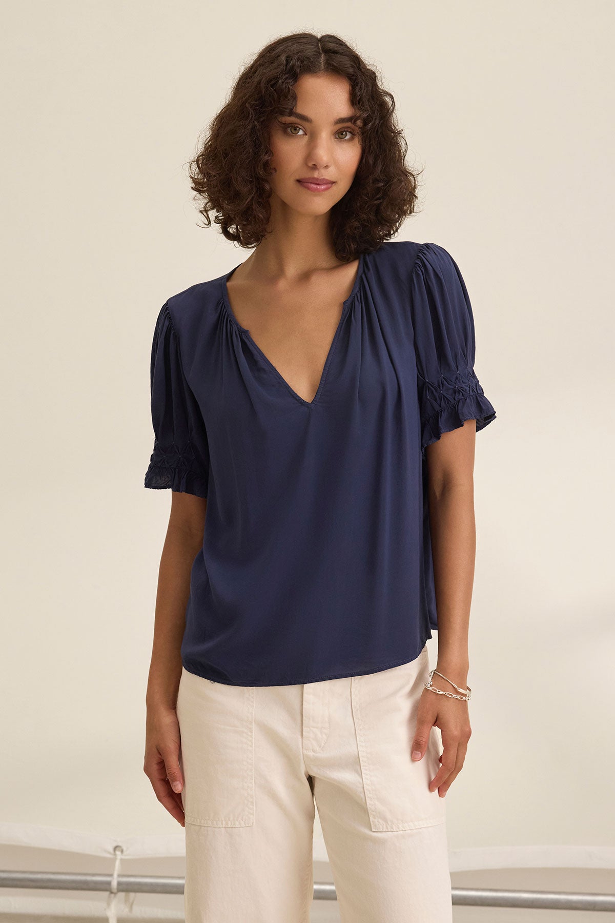 A woman with curly hair wears the Velvet by Graham & Spencer CALISSA SPLIT NECK BLOUSE, a chic navy V-neck top with puffed sleeves, paired with high-waisted cream pants against a neutral background.