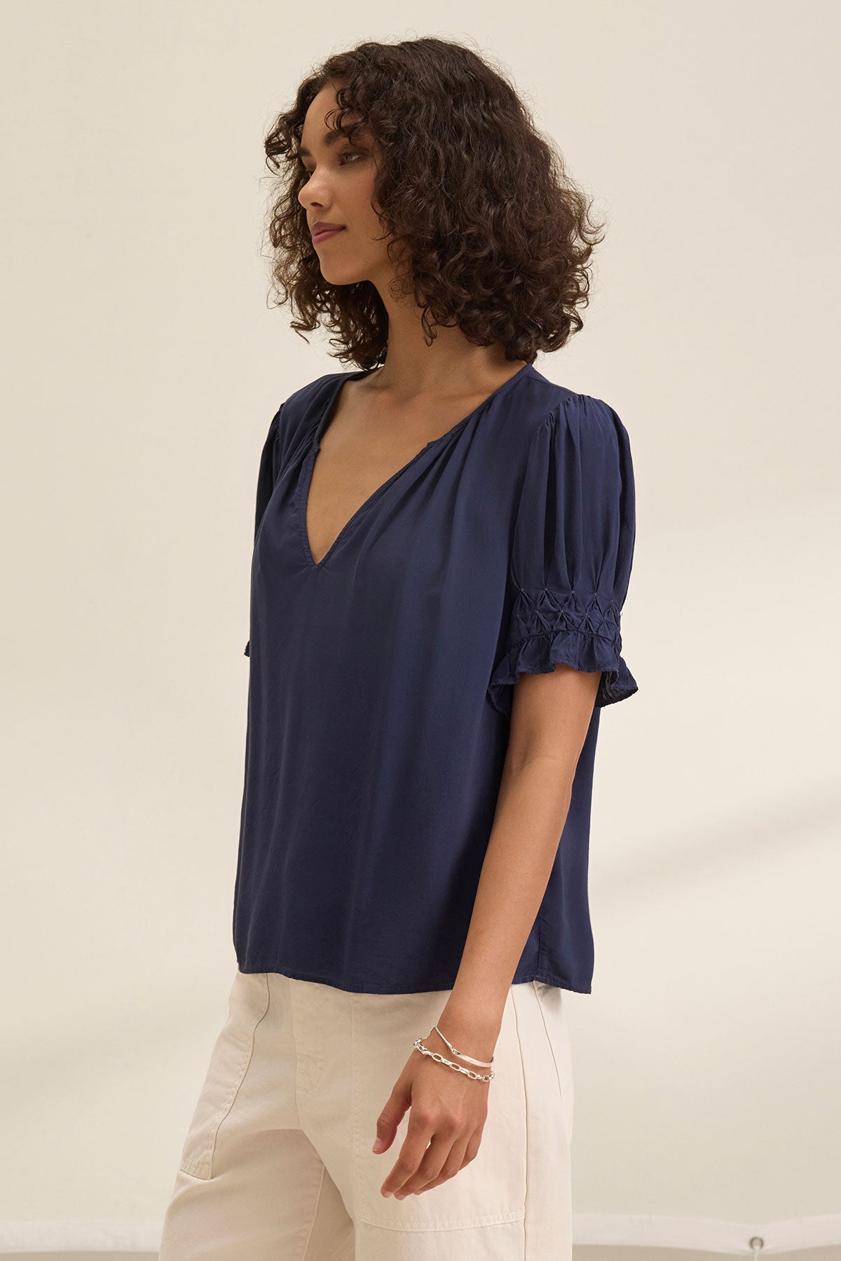 A person with curly hair wears the CALISSA SPLIT NECK BLOUSE by Velvet by Graham & Spencer—a relaxed fit, navy blue women's rayon challis top with short sleeves and a V-neck—paired with off-white pants against a plain light background.