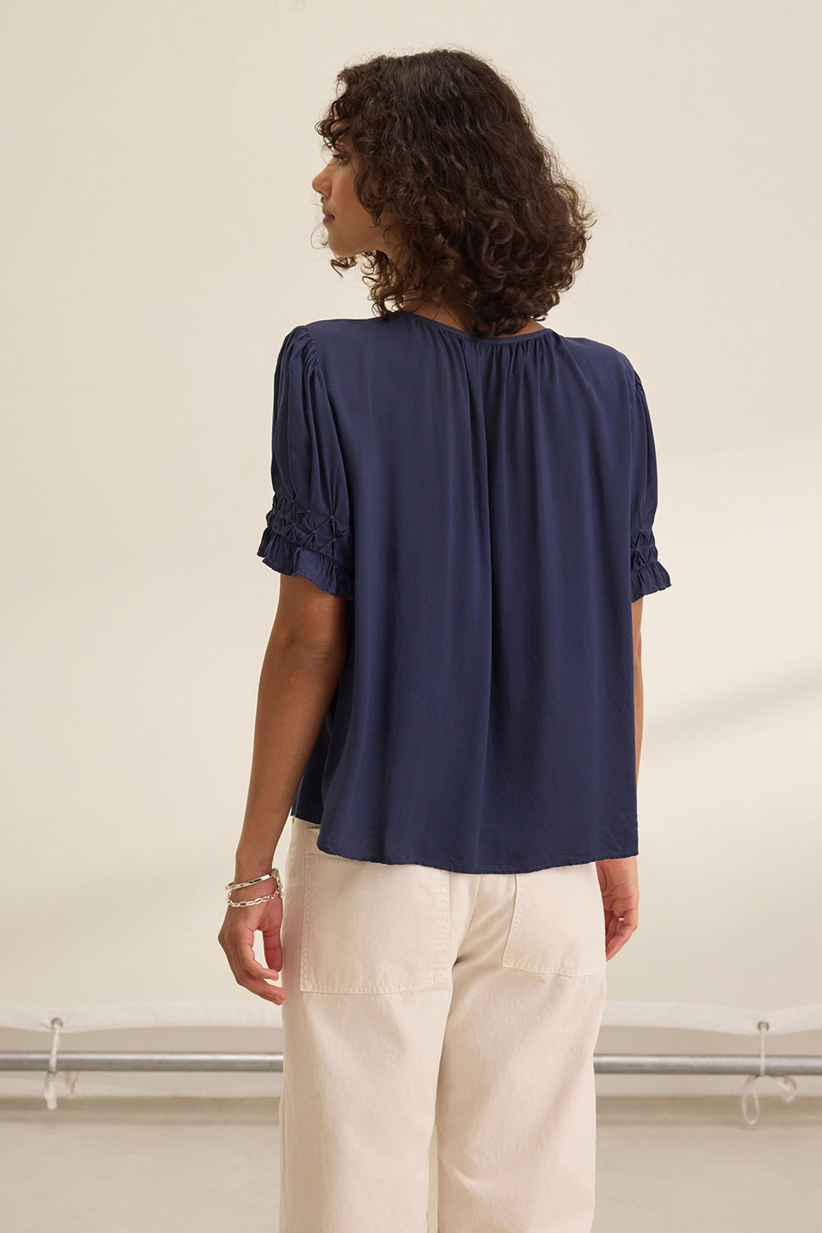 A person with curly hair stands with their back to the camera, wearing the Velvet by Graham & Spencer CALISSA SPLIT NECK BLOUSE in navy blue and light-colored pants in a neutral indoor setting.