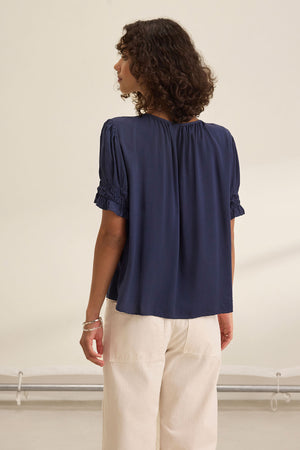 A person with curly hair stands with their back to the camera, wearing the Velvet by Graham & Spencer CALISSA SPLIT NECK BLOUSE in navy blue and light-colored pants in a neutral indoor setting.