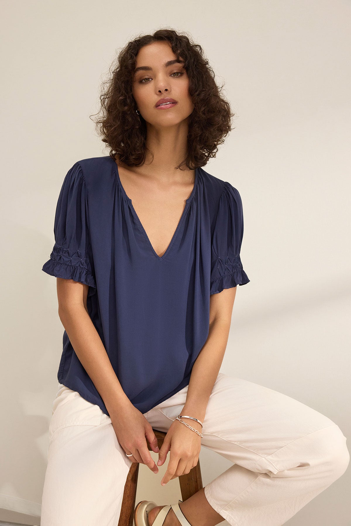 A woman with curly hair wears the CALISSA SPLIT NECK BLOUSE in navy rayon challis by Velvet by Graham & Spencer and off-white pants, sitting on a stool against a plain light background.