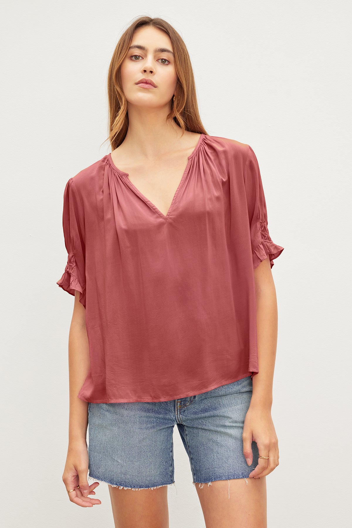A person with long hair wears a loose-fitting, short-sleeved, deep pink CALISSA SPLIT NECK BLOUSE by Velvet by Graham & Spencer with a relaxed fit and denim shorts.-36909459144897