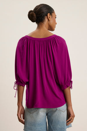 A person faces away, wearing the LUNA TOP by Velvet by Graham & Spencer—a bright magenta rayon challis blouse with gathered sleeves—paired with light blue denim shorts, set against a neutral background.