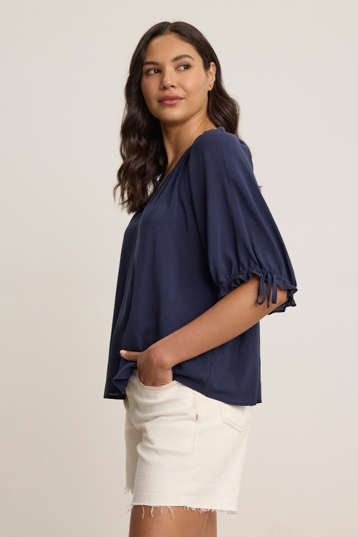 A woman stands sideways against a plain light background, wearing the versatile LUNA TOP by Velvet by Graham & Spencer in soft navy Rayon Challis with elbow-length sleeves, paired with white frayed hem shorts and one hand in her pocket.