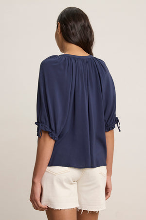 A woman with long dark hair, seen from the back, wears the Velvet by Graham & Spencer LUNA TOP— a versatile navy Rayon Challis V-neck with elbow-length sleeves—paired with white frayed hem shorts.