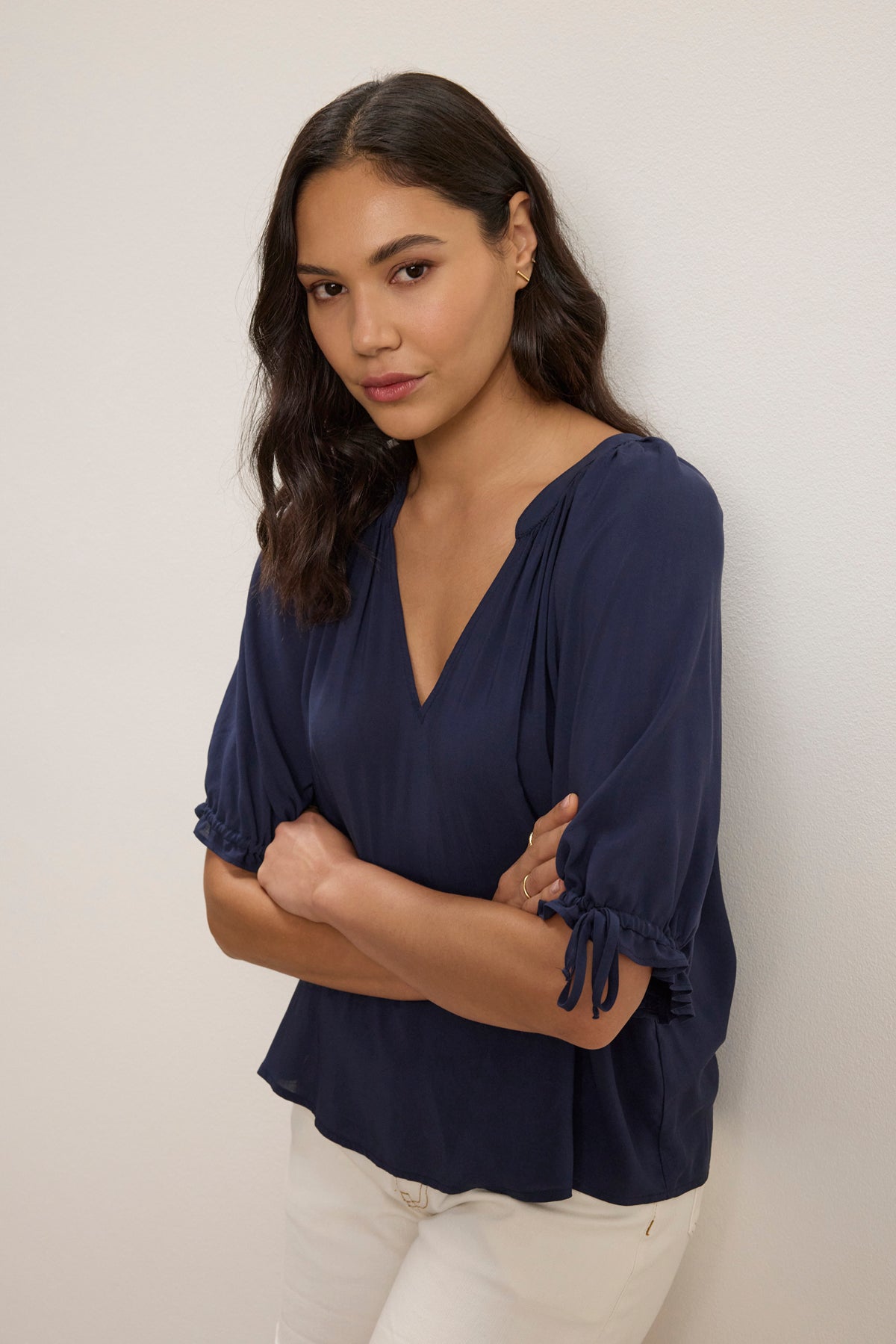 A woman with medium-length dark hair wears the Velvet by Graham & Spencer LUNA TOP—a versatile navy V-neck, relaxed-fit top with gathered sleeves—paired with white pants, standing against a light wall with arms crossed.