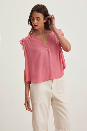 A woman in the Velvet by Graham & Spencer MALTA TOP, featuring pink flutter sleeves and a v-neckline, stands with one hand touching her hair against a plain background, showcasing this versatile top's effortless style paired with white pants.