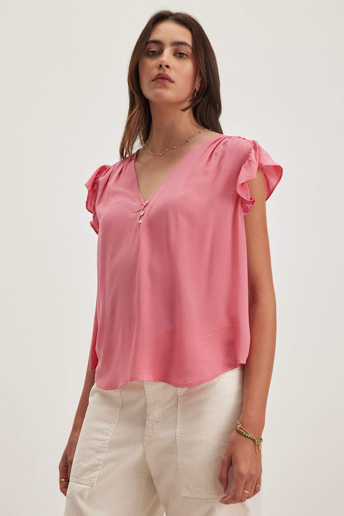 A woman models the Velvet by Graham & Spencer MALTA TOP, a versatile pink V-neck blouse with flutter sleeves, paired with white pants against a neutral background.