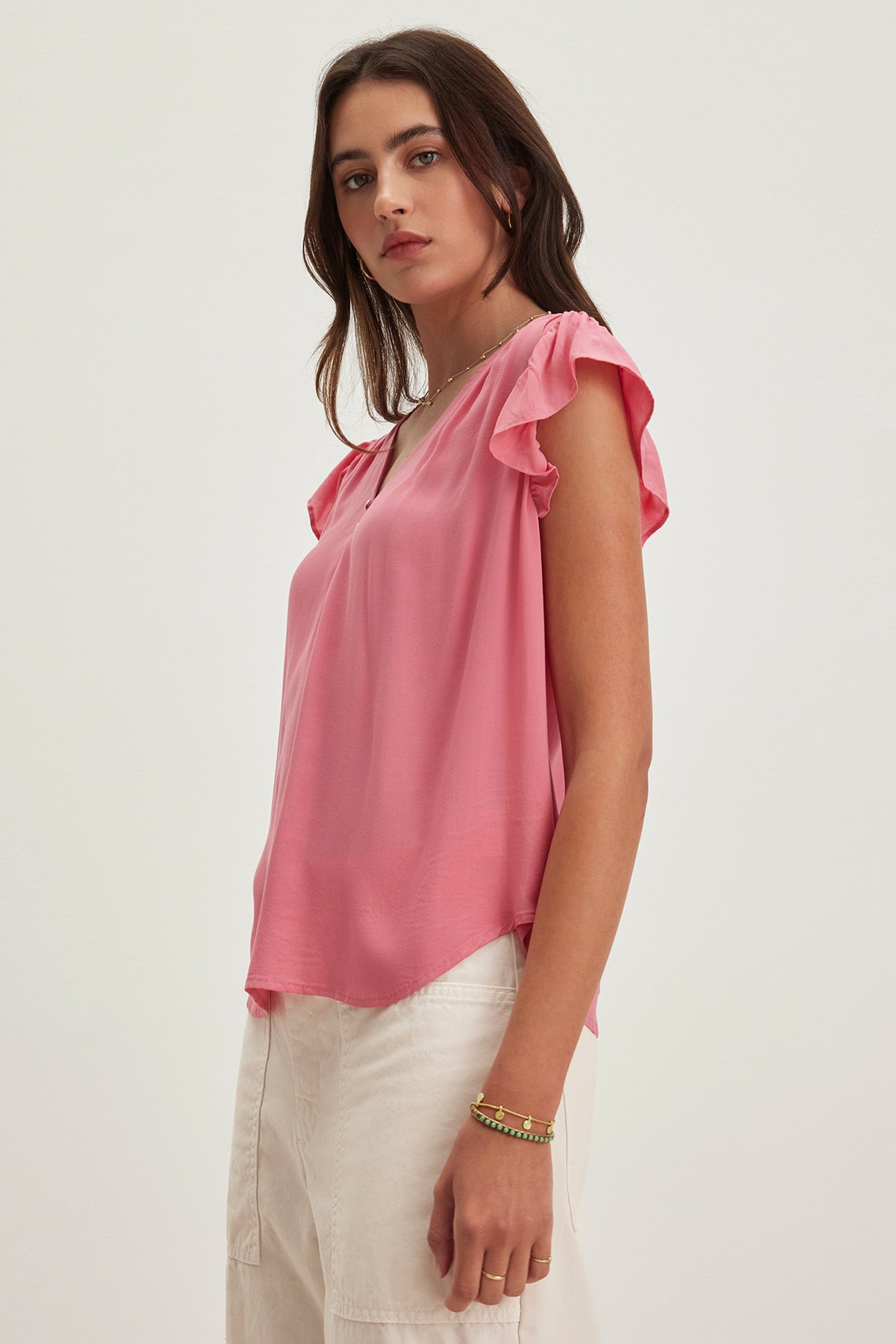 A woman wearing the MALTA TOP by Velvet by Graham & Spencer, featuring a pink ruffled sleeveless design with a V-neckline, paired with cream pants, gazes at the camera against a neutral background.