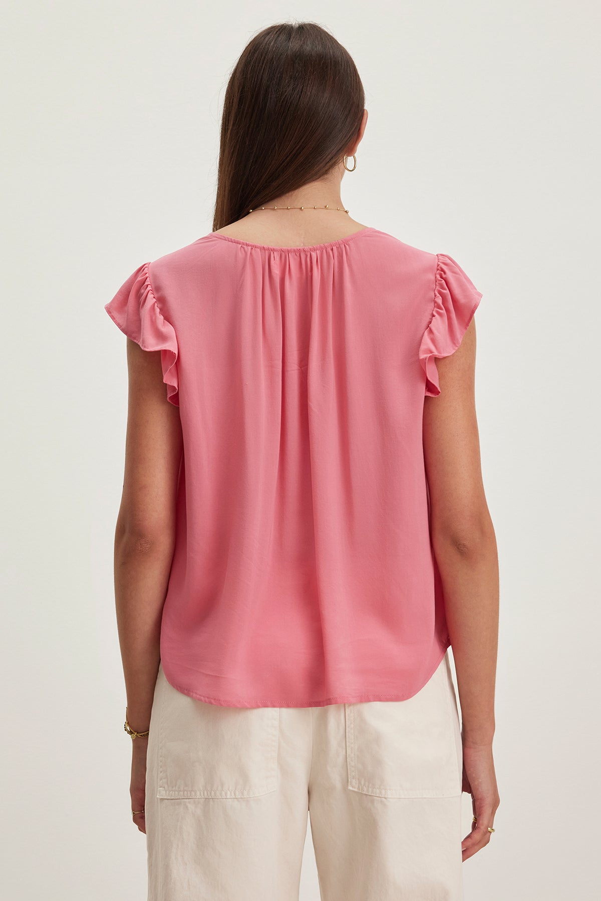 A person with long hair is seen from the back, wearing a Velvet by Graham & Spencer MALTA TOP in pink with flutter sleeves and white pants.