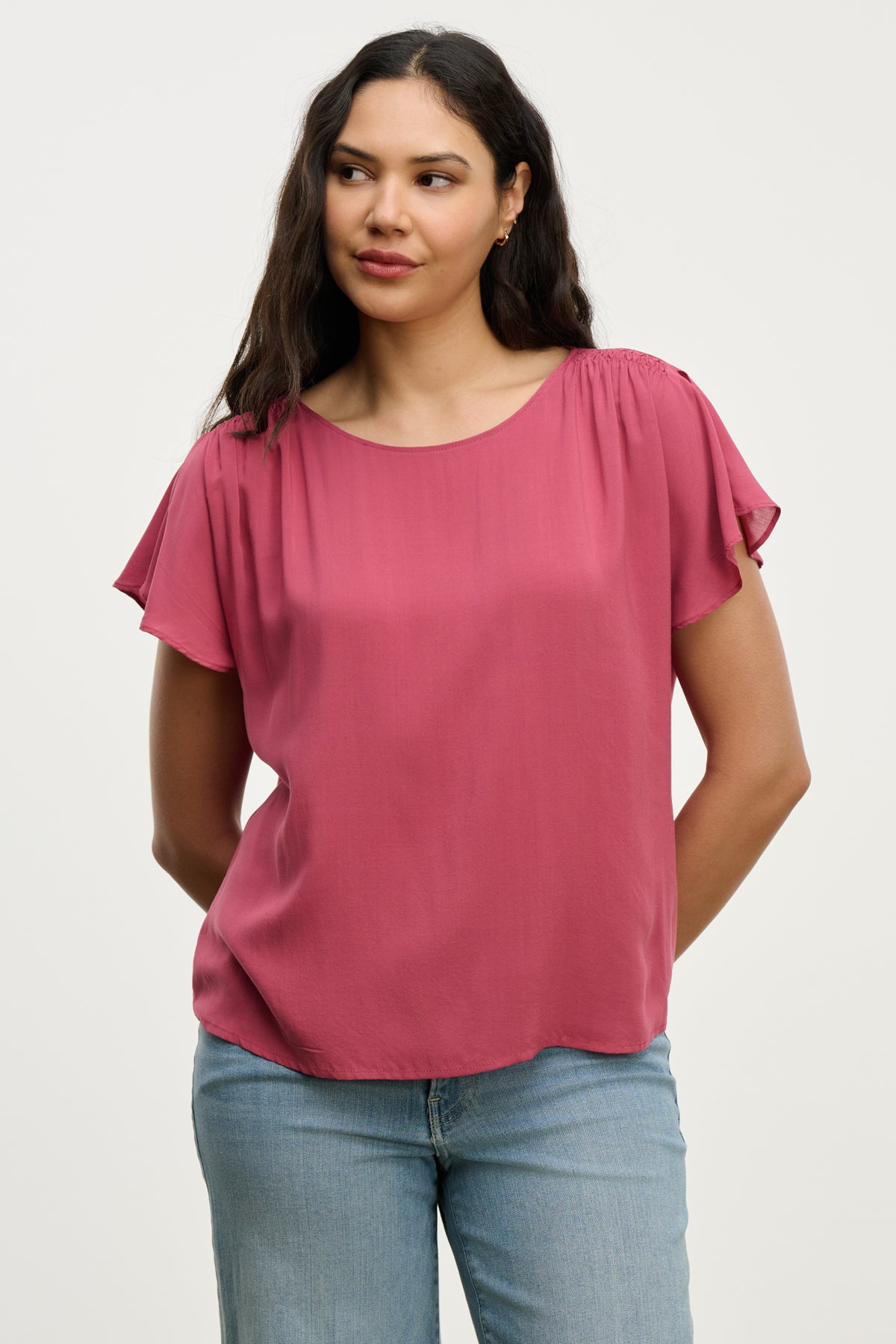 A woman wears the MARCEL TOP by Velvet by Graham & Spencer— a short-sleeve, rose-colored rayon challis blouse with smocked shoulders—paired with light blue jeans, standing against a plain light background.
