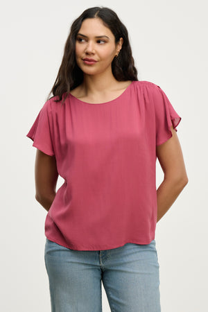 A woman wears the MARCEL TOP by Velvet by Graham & Spencer— a short-sleeve, rose-colored rayon challis blouse with smocked shoulders—paired with light blue jeans, standing against a plain light background.