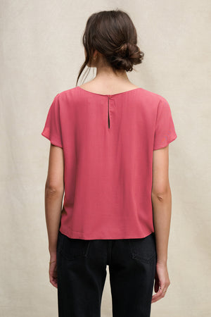 A woman with brown hair in a loose bun wears the Velvet by Graham & Spencer MARCEL TOP, a short-sleeved pink rayon challis top with a keyhole closure, and black pants, standing with her back to the camera against a neutral background.