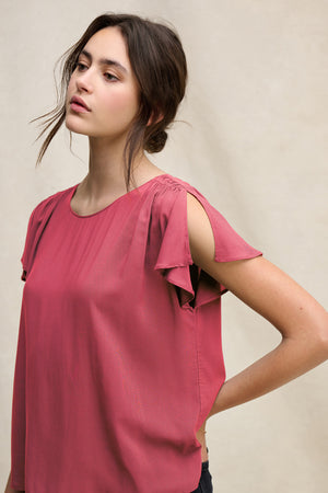 A woman with long brown hair in a loose bun wears the MARCEL TOP by Velvet by Graham & Spencer, a rose-colored rayon challis blouse with split shoulder details and a keyhole closure, standing against a neutral background.