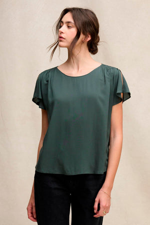 A woman wears the MARCEL TOP by Velvet by Graham & Spencer, featuring a round neckline, short sleeves, and smocked shoulders, paired with black pants against a plain light background.