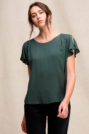 A woman wears the MARCEL TOP by Velvet by Graham & Spencer—a dark green rayon challis top with a round neckline and shoulder slits—paired with black pants, standing against a neutral background.