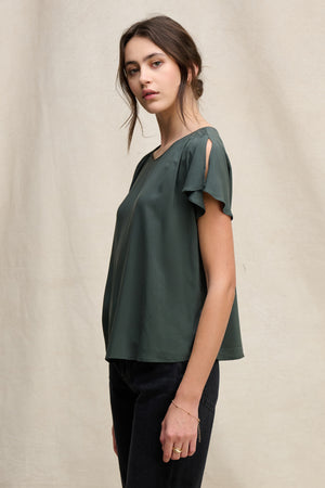 A woman stands sideways in the MARCEL TOP by Velvet by Graham & Spencer—dark green rayon challis with smocked shoulders—paired with black pants, posing against a plain beige backdrop.