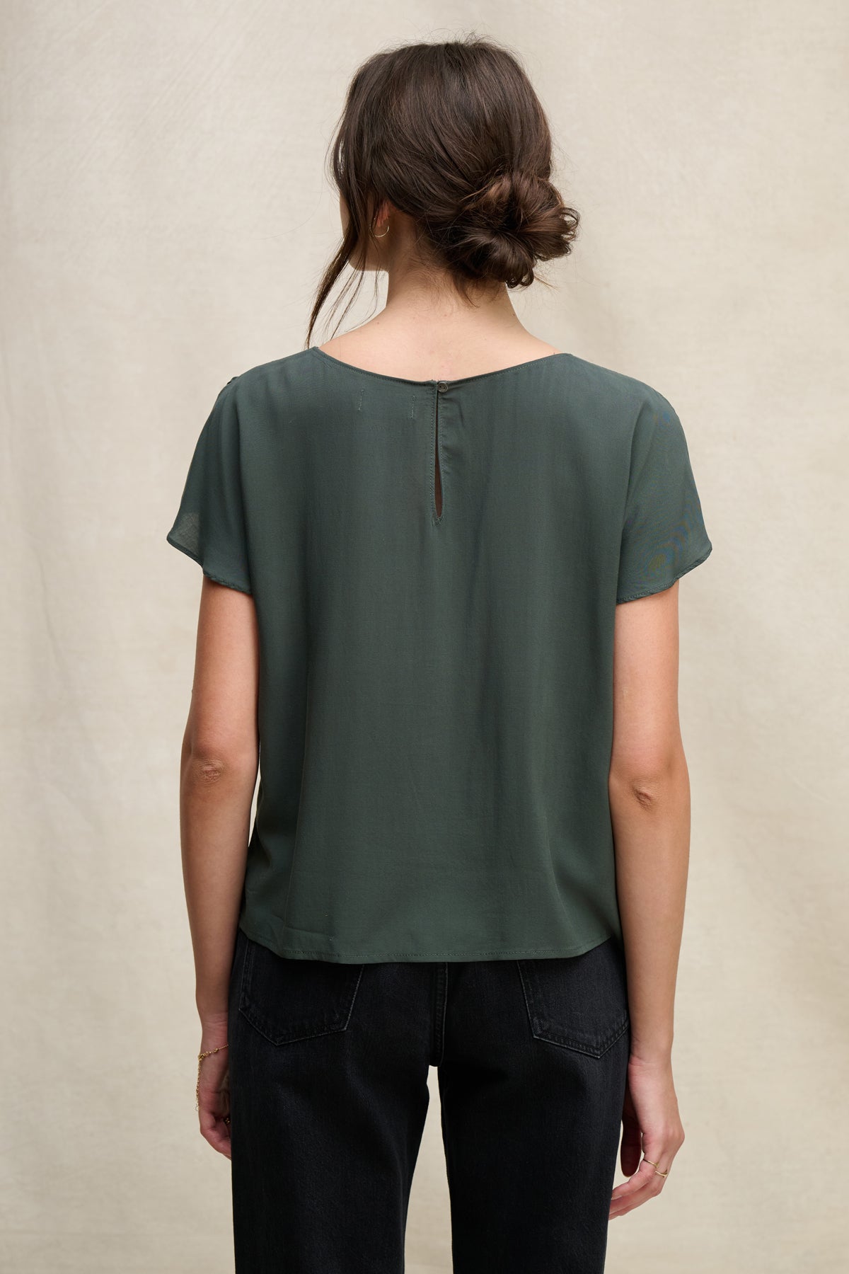 A woman with brown hair in a loose bun is seen from the back, wearing Velvet by Graham & Spencer’s MARCEL TOP in dark green with a round neckline and black jeans, posed against a neutral background.