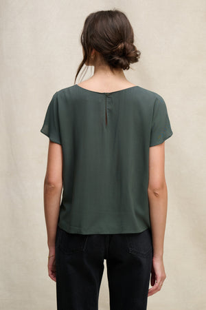 A woman with brown hair in a loose bun is seen from the back, wearing Velvet by Graham & Spencer’s MARCEL TOP in dark green with a round neckline and black jeans, posed against a neutral background.