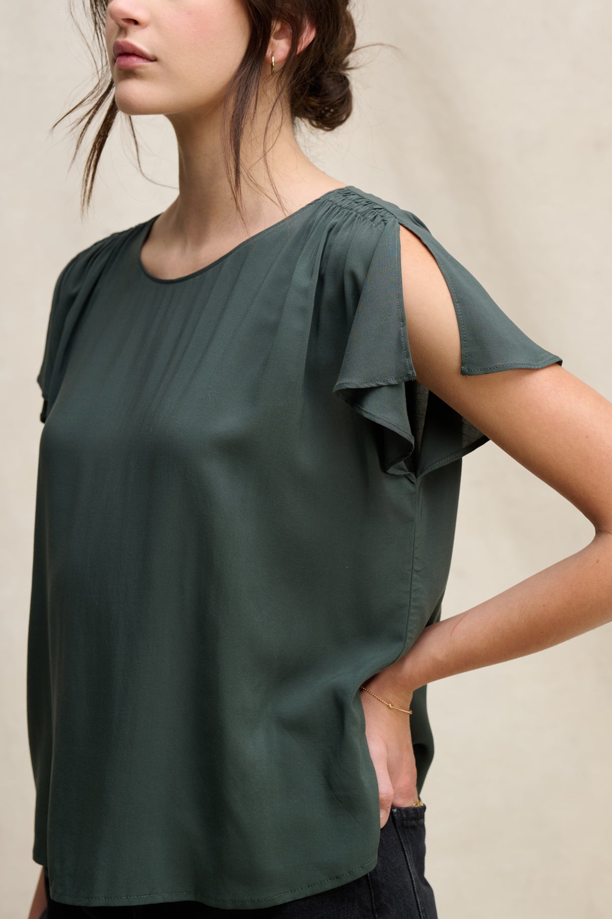 A woman models the MARCEL TOP by Velvet by Graham & Spencer—a dark green rayon challis top featuring pleated details, round neckline, and sleeve slits—as she stands with one hand on her hip against a neutral background.
