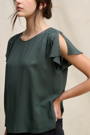 A woman models the MARCEL TOP by Velvet by Graham & Spencer—a dark green rayon challis top featuring pleated details, round neckline, and sleeve slits—as she stands with one hand on her hip against a neutral background.