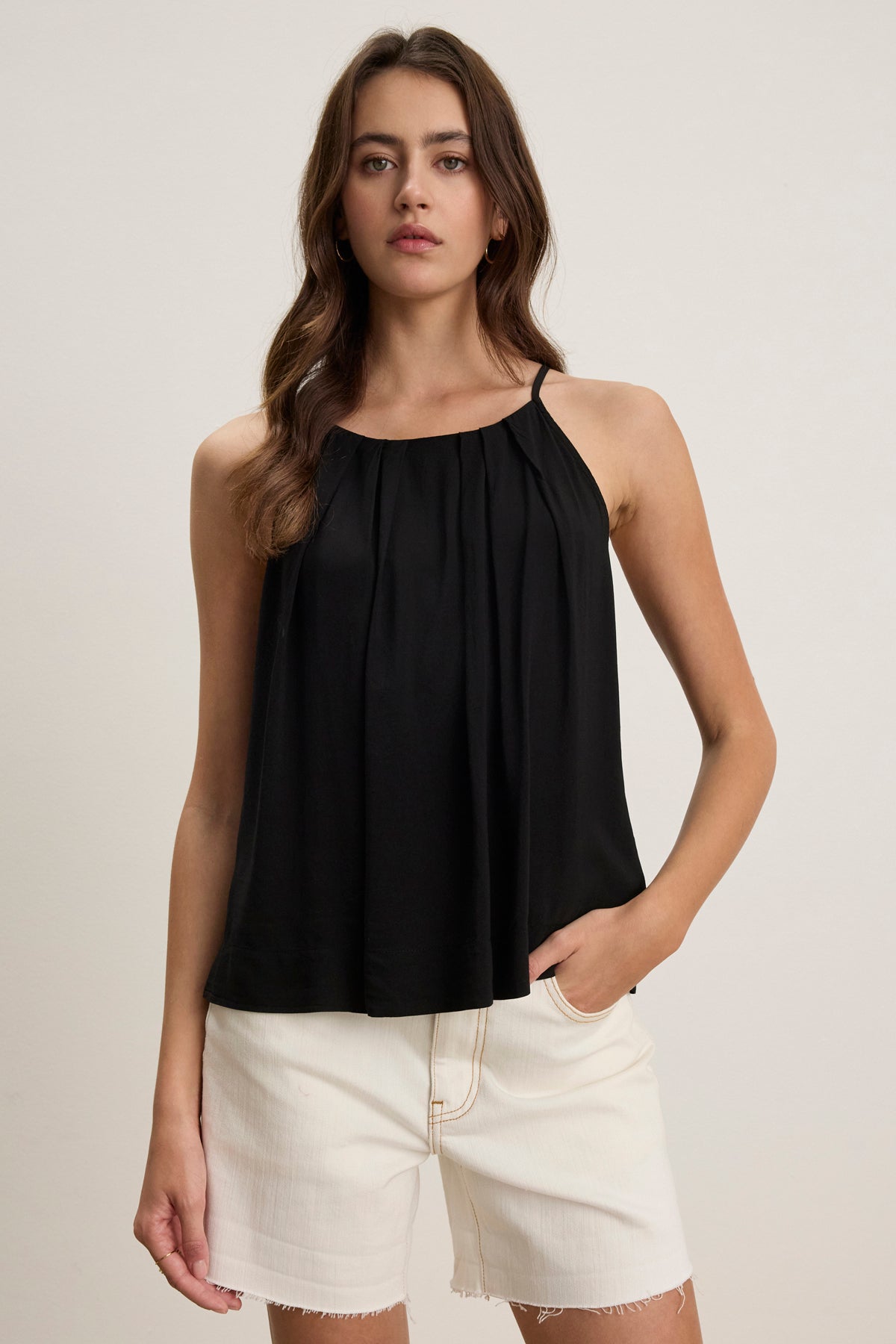 A woman wears the Velvet by Graham & Spencer PIPER TANK TOP, a sleeveless black Rayon Challis high-neck tank, paired with white shorts, standing against a plain light background.