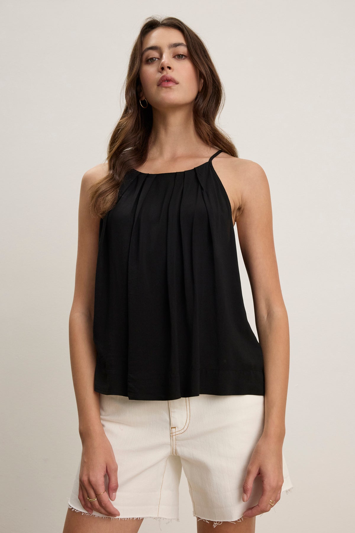 A woman models the PIPER TANK TOP by Velvet by Graham & Spencer, a sleeveless black pleated high neck style, paired with white frayed hem shorts against a plain light background.