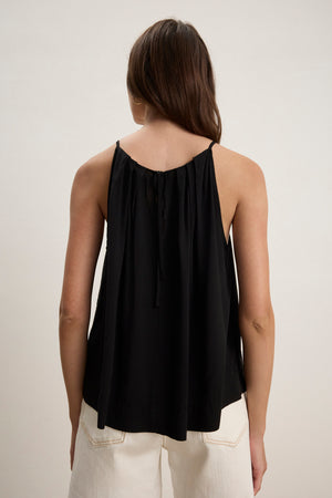 A woman with long brown hair faces away, wearing the Velvet by Graham & Spencer PIPER TANK TOP—a black, pleated high-neck tank with thin straps—paired with white pants against a plain background.