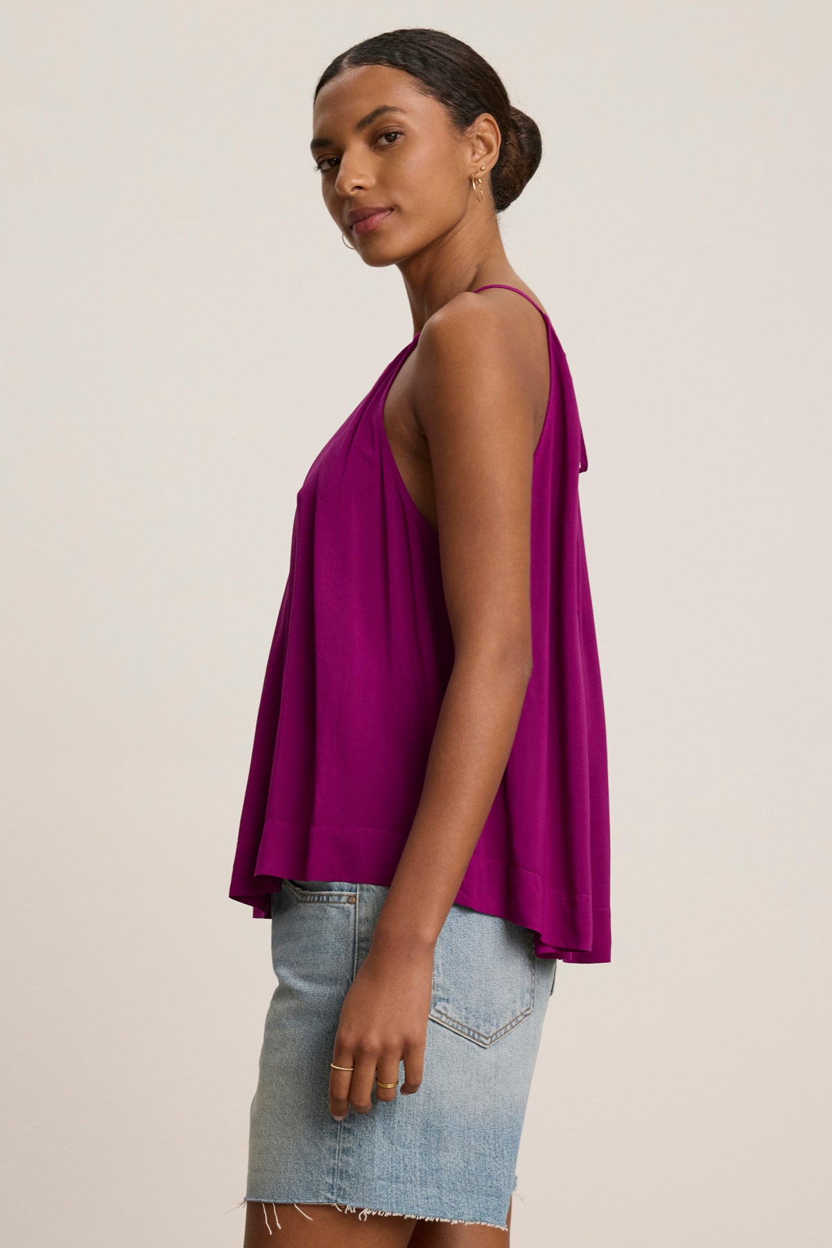 A woman in profile wears the Velvet by Graham & Spencer PIPER TANK TOP, a loose-fitting, sleeveless, high neck magenta top in Rayon Challis, paired with a light blue denim skirt against a plain background.