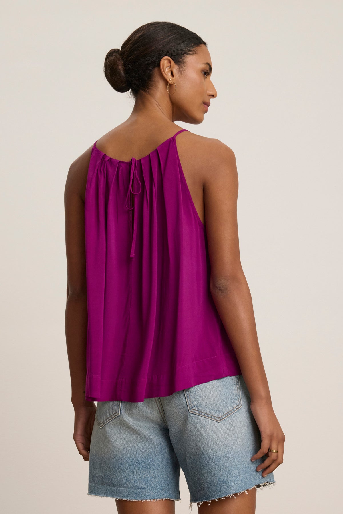 A woman stands facing away from the camera, wearing the sleeveless magenta PIPER TANK TOP by Velvet by Graham & Spencer—featuring a high neck, tied back, and pleated fabric—paired with light blue denim shorts.