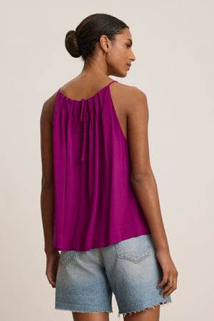 A woman stands facing away from the camera, wearing the sleeveless magenta PIPER TANK TOP by Velvet by Graham & Spencer—featuring a high neck, tied back, and pleated fabric—paired with light blue denim shorts.
