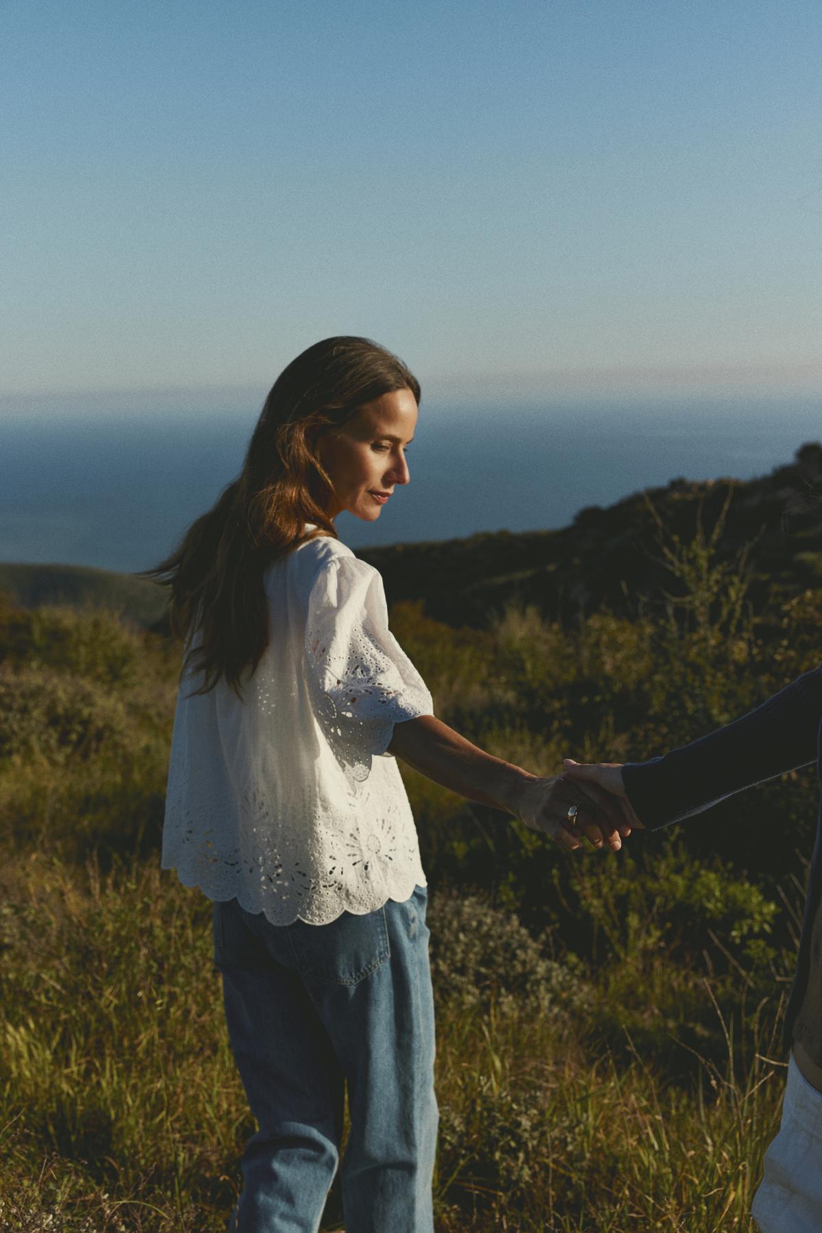 A woman in a RAZI EMBROIDERED COTTON LACE TOP by Velvet by Graham & Spencer, and blue jeans holding hands with someone off-camera, standing in a sunny, grassy hillside landscape.-36443805712577
