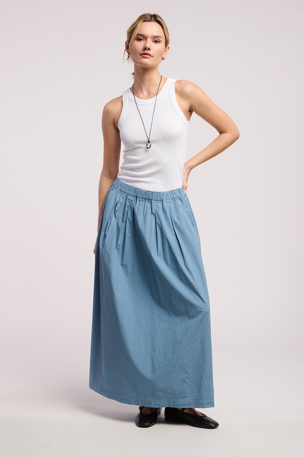 A person stands against a plain background, wearing a white sleeveless top, the CHESSA SKIRT by Velvet by Graham & Spencer—a cotton poplin A-line maxi skirt with an elastic waist—paired with black shoes and a necklace.-41645003505857