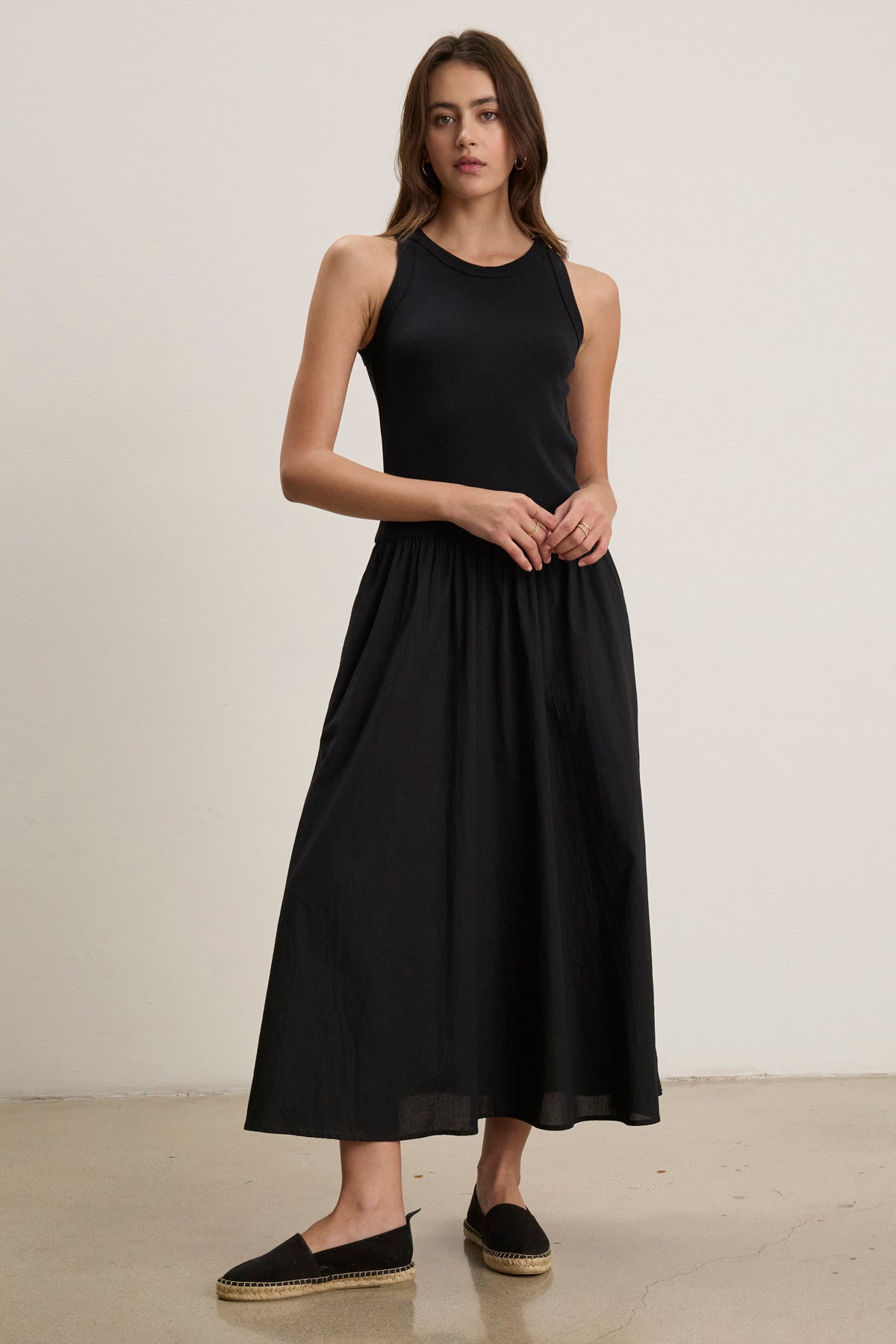 A woman wearing the sleeveless AMELIA DRESS by Velvet by Graham & Spencer, made of black cotton poplin with side seam pockets, and black espadrilles stands against a plain light-colored wall on a polished floor.-39111854981313