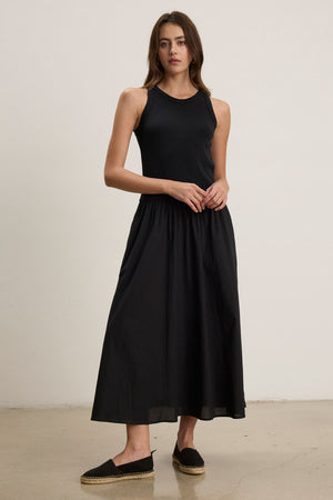 A woman wearing the sleeveless AMELIA DRESS by Velvet by Graham & Spencer, made of black cotton poplin with side seam pockets, and black espadrilles stands against a plain light-colored wall on a polished floor.