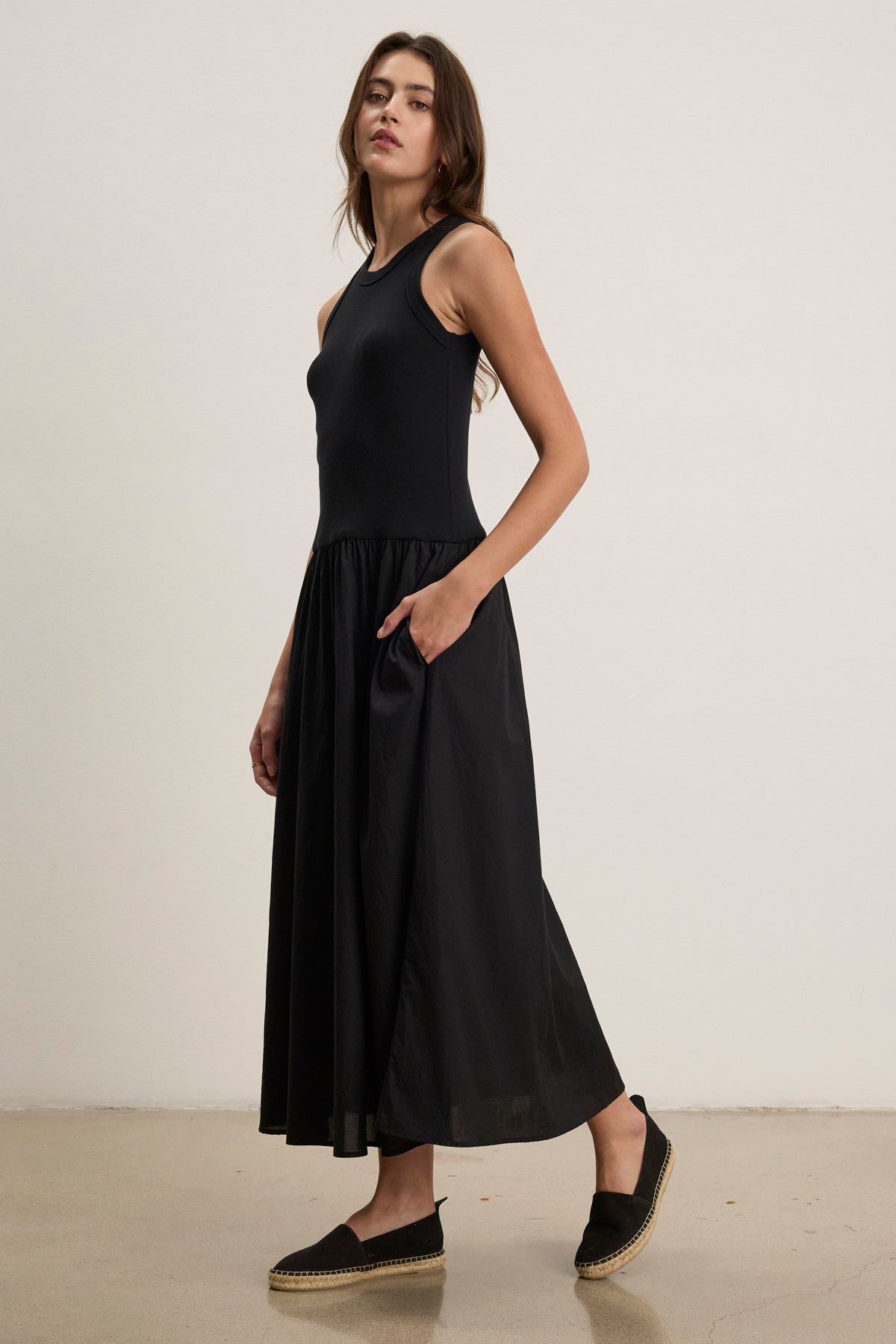A woman stands against a plain wall, wearing the Velvet by Graham & Spencer AMELIA DRESS— a sleeveless black cotton poplin dress with a flared skirt and side seam pockets—paired with black slip-on shoes, gazing slightly to the side.-39111855014081