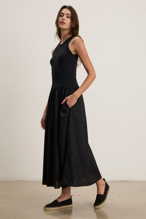 A woman stands against a plain wall, wearing the Velvet by Graham & Spencer AMELIA DRESS— a sleeveless black cotton poplin dress with a flared skirt and side seam pockets—paired with black slip-on shoes, gazing slightly to the side.