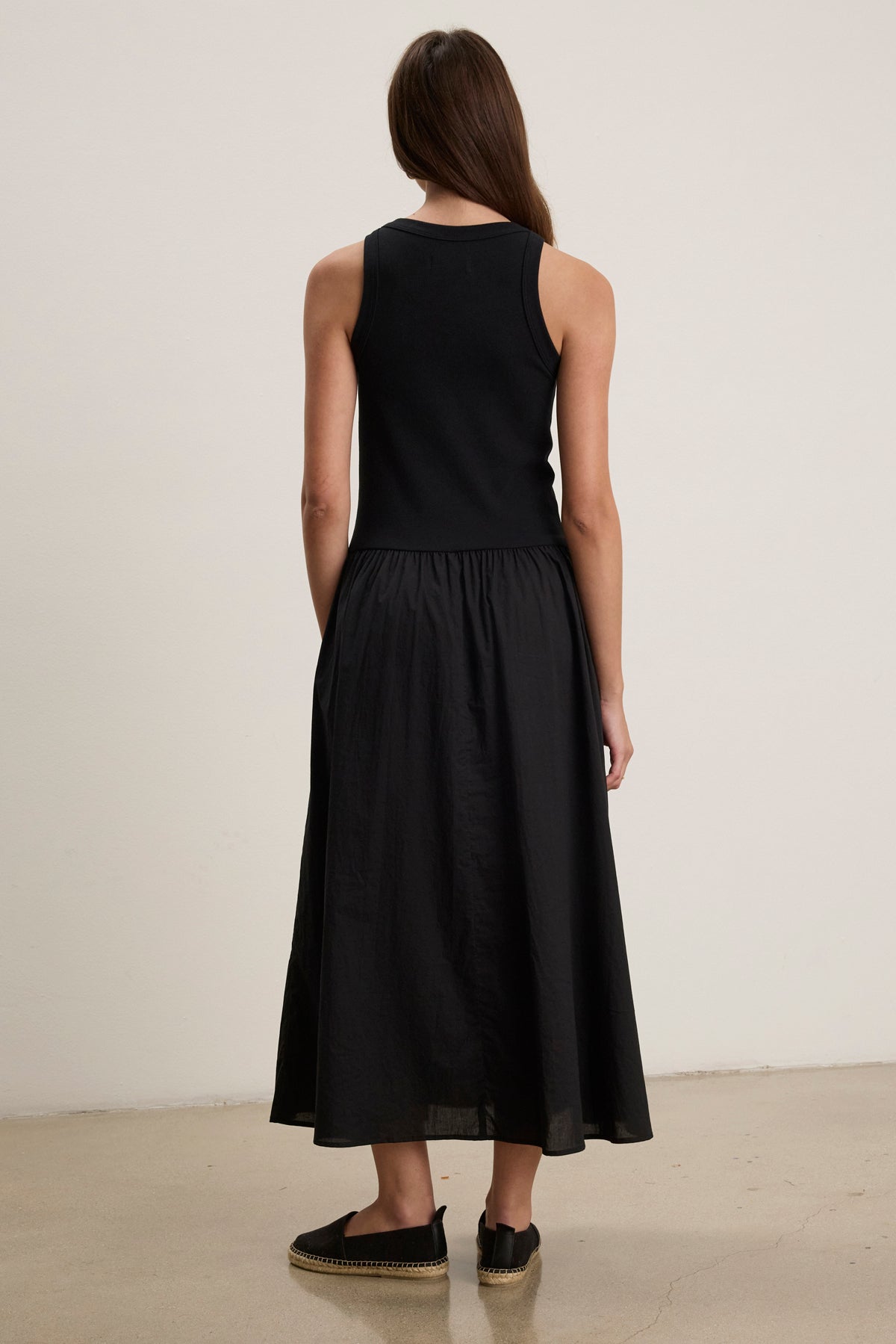 A person stands indoors with their back to the camera, wearing the AMELIA DRESS by Velvet by Graham & Spencer—a sleeveless black cotton poplin dress with side seam pockets—paired with black slip-on shoes.-39111855079617