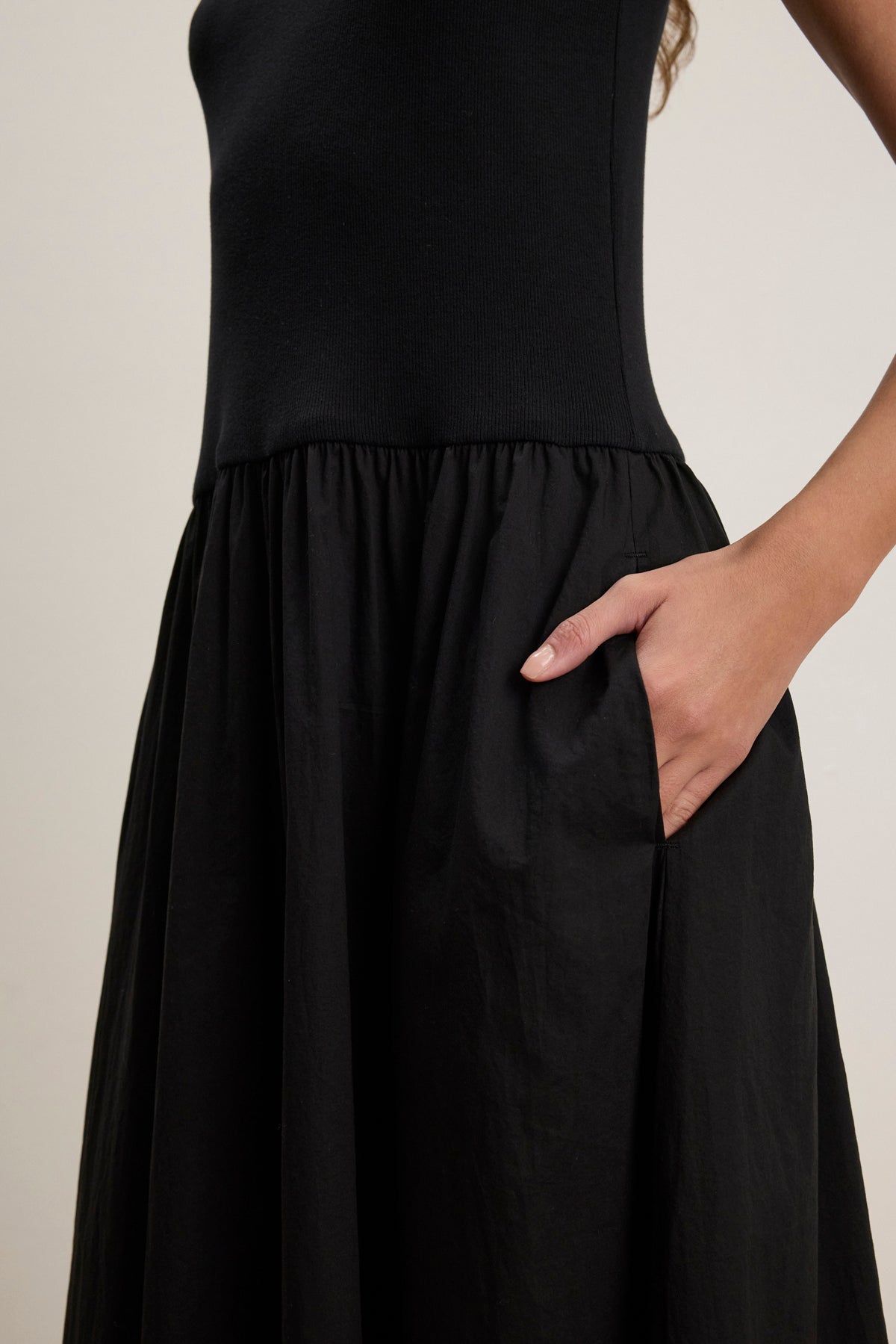A person stands with one hand in a side pocket, wearing the AMELIA DRESS by Velvet by Graham & Spencer—a sleeveless black dress featuring a gathered skirt and crafted from stretchy ribbed cotton.-39111855177921