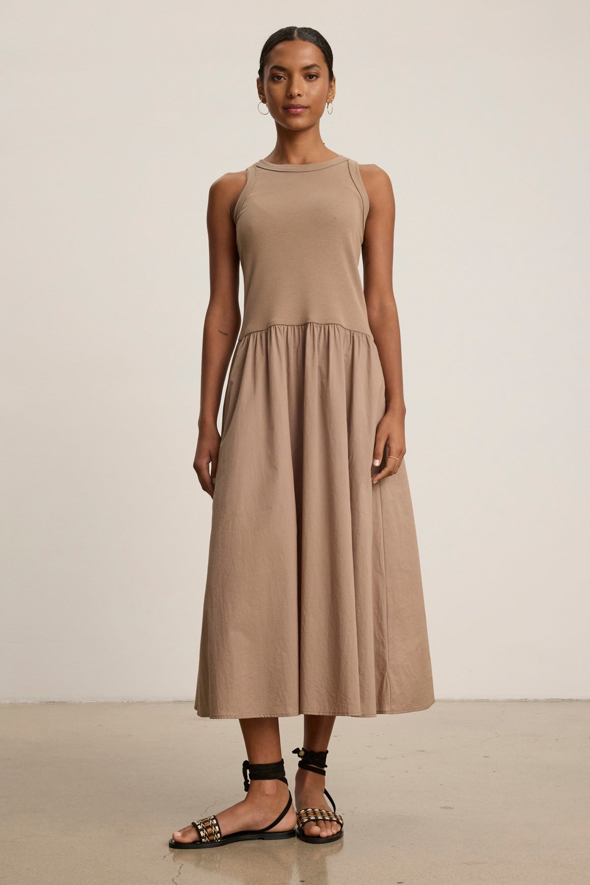 A woman poses against a plain background in the AMELIA DRESS by Velvet by Graham & Spencer, a sleeveless, beige, ankle-length cotton-elastane dress, styled with black studded sandals.