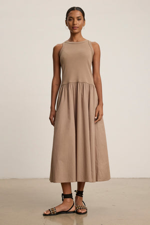 A woman poses against a plain background in the AMELIA DRESS by Velvet by Graham & Spencer, a sleeveless, beige, ankle-length cotton-elastane dress, styled with black studded sandals.