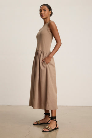 A woman stands in profile against a plain background wearing Velvet by Graham & Spencer’s AMELIA DRESS, a versatile taupe ankle-length style made from stretchy ribbed cotton with pockets, paired with black strappy sandals.