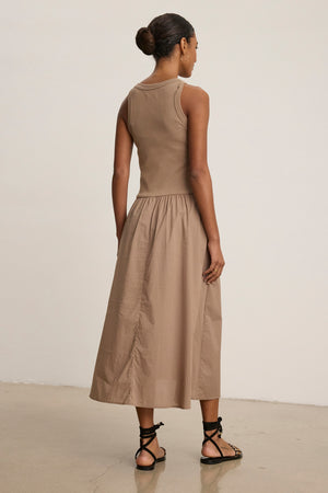 A woman stands indoors facing away, wearing the AMELIA DRESS by Velvet by Graham & Spencer—a ribbed beige cotton-elastane sleeveless midi dress—paired with black strappy sandals.