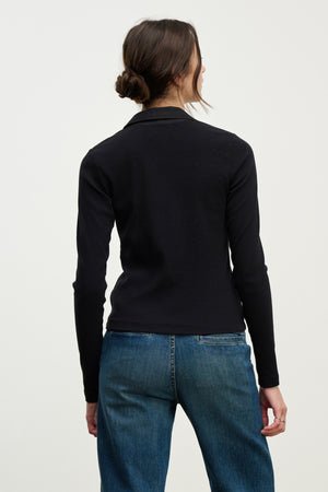 A woman with brown hair in a low bun wears the ARVINE SHIRT by Velvet by Graham & Spencer, a black ribbed fitted top, paired with blue jeans, standing with her back to a plain light background.