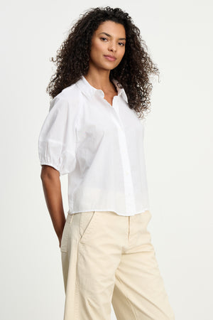 A woman with curly hair wears the AUTUMN SHIRT by Velvet by Graham & Spencer and light beige pants, standing against a plain light background—perfect for effortless everyday style.