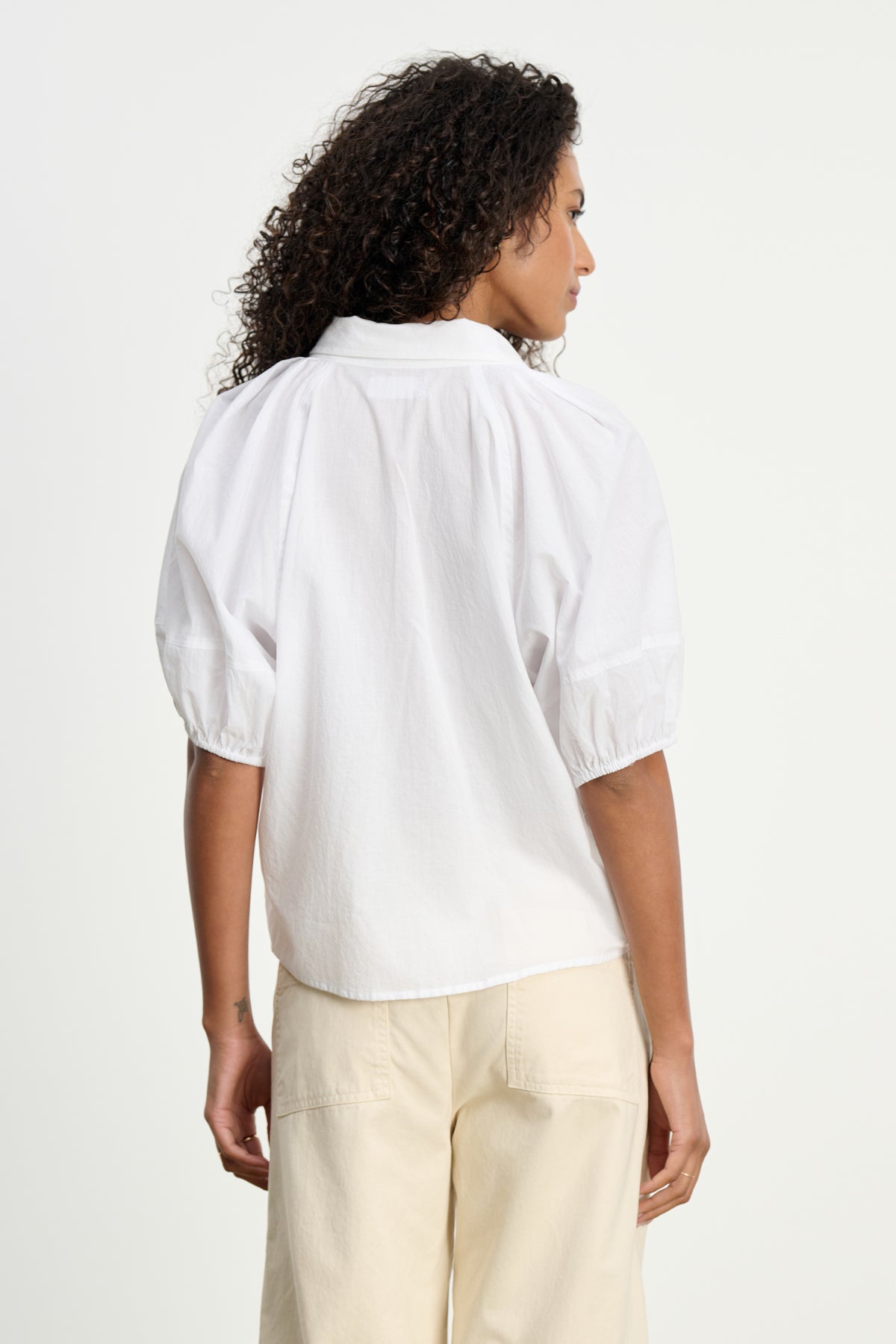 A woman with curly hair is seen from the back in the AUTUMN SHIRT by Velvet by Graham & Spencer and light beige pants, set against a plain backdrop—a chic choice for everyday style.