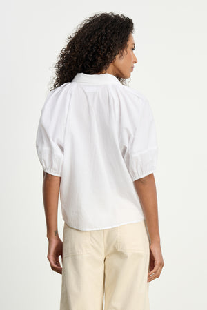 A woman with curly hair is seen from the back in the AUTUMN SHIRT by Velvet by Graham & Spencer and light beige pants, set against a plain backdrop—a chic choice for everyday style.