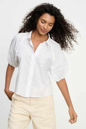 A woman wears the AUTUMN SHIRT by Velvet by Graham & Spencer, a white cotton poplin top with puff sleeves, paired with beige pants and standing against a plain light background.